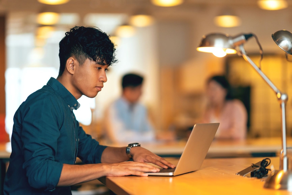 A man sits at a desk working on a laptop. In the background, colleagues are talking.